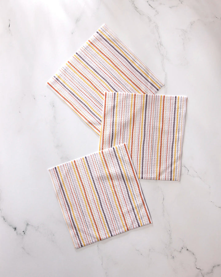 Three GEOMETRY Stripe Season white napkins with vertical multicolored stripes, made from recycled materials, are neatly arranged and slightly overlapping on a white marble surface for a stylish, sustainable touch.