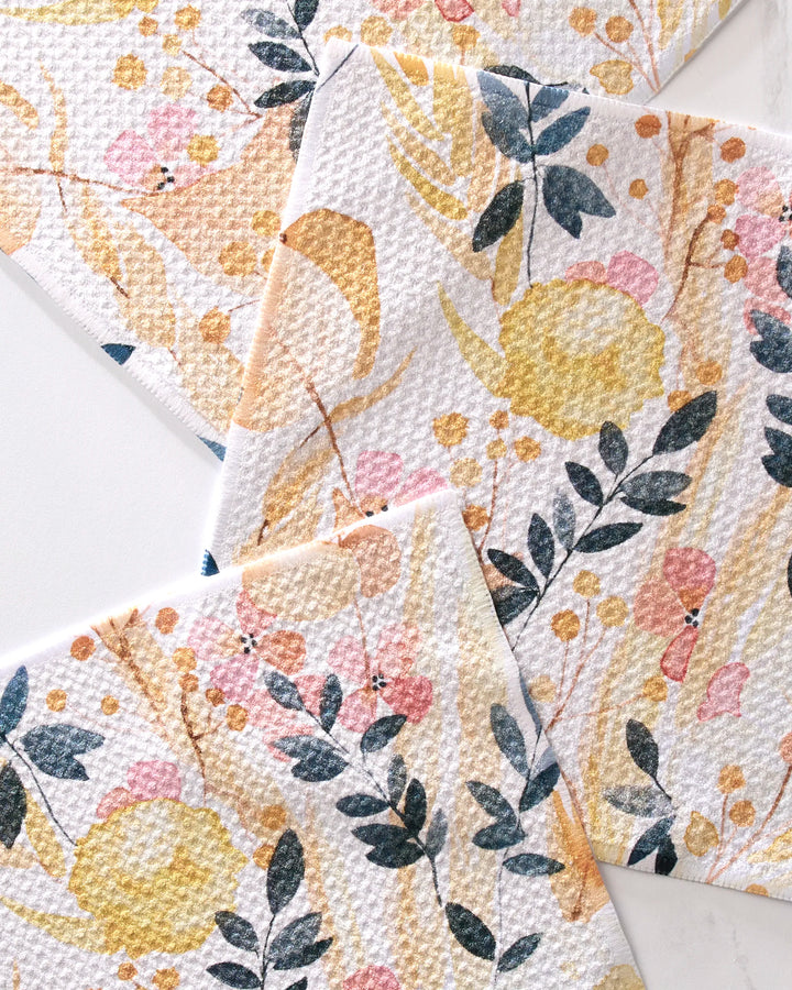 Three GEOMETRY Sleepy Fields kitchen towels with a textured surface and floral print in yellow, pink, black, and gray are overlapped on a light background.