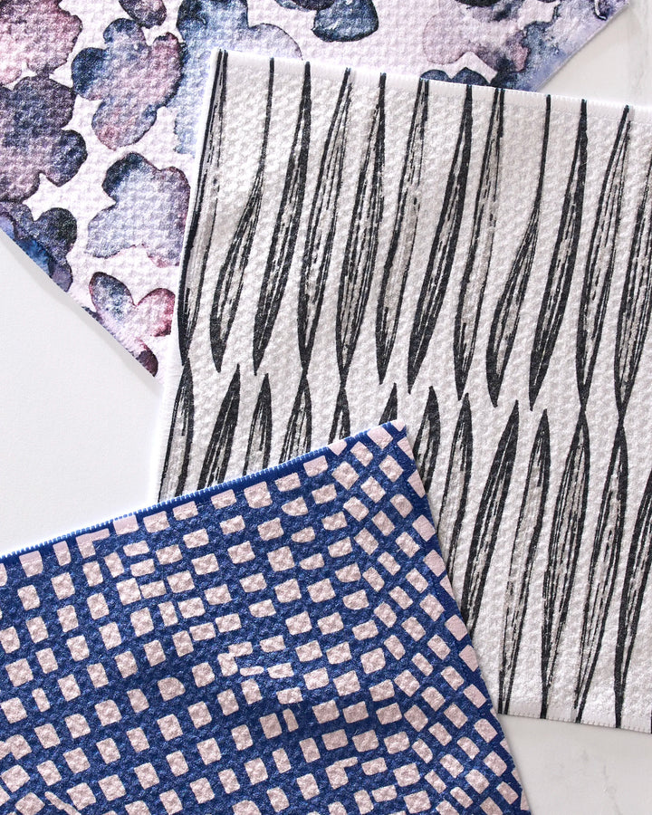 Three pieces from GEOMETRY’s Tiny Petals set—a purple watercolor-spotted, a black leaf-patterned, and a blue-and-white geometric towel—overlap on a white surface. These super-absorbent towels are made from recycled materials.