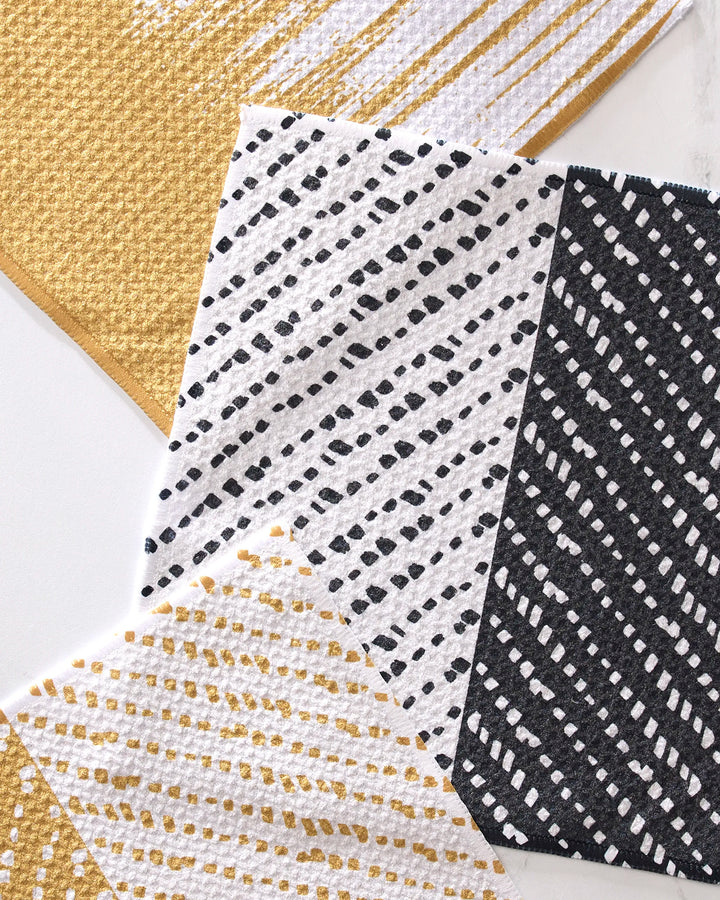 Three GEOMETRY This And That recycled dishcloths overlap on marble. Patterns include gold and white dots, black and white dots, or white with gold dots. One absorbent cloth features gold diagonal stripes along the edge for accent.