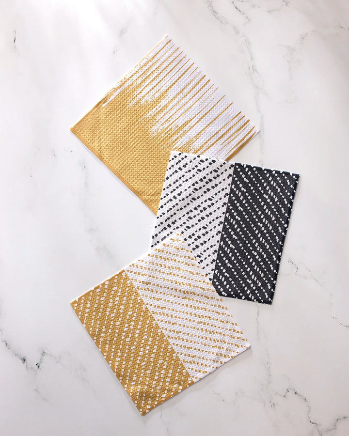 Three square GEOMETRY "This And That" dishcloths, featuring absorbent fabric and diagonal patterns in gold and white, black and white, plus a solid textured gold design, are arranged on a white marble surface.