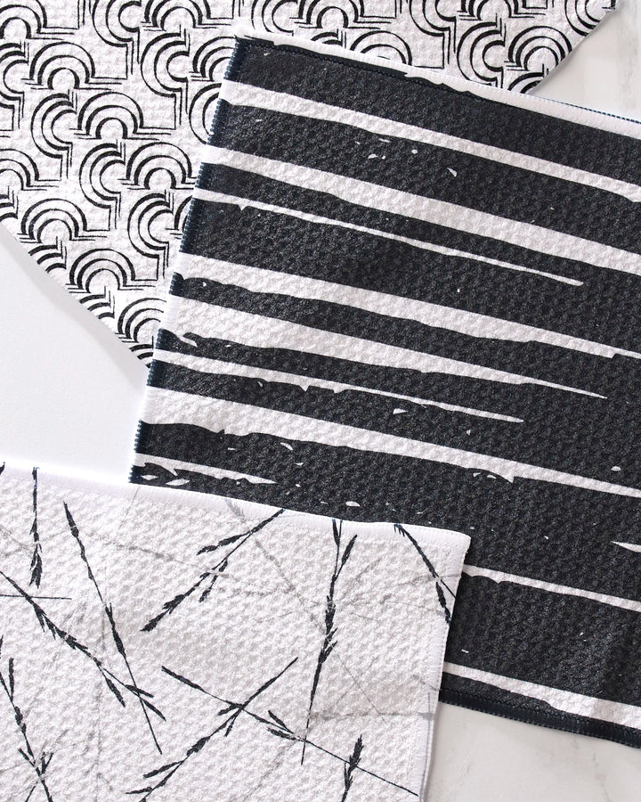 The GEOMETRY Night Reeds dishcloth set features three eco-friendly cloths in black and white rainbow, stripe, and abstract patterns, arranged diagonally on a white surface. Made from recycled materials for stylish, sustainable cleaning.