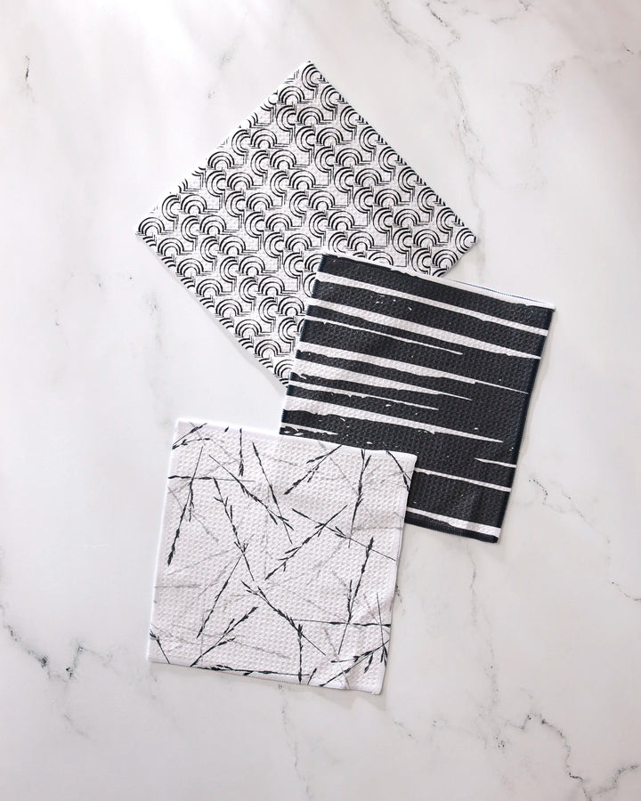 The GEOMETRY Night Reeds dishcloth set includes three square cloths with black and white geometric patterns—scallops, stripes, and intersecting lines—displayed on a white marble surface.