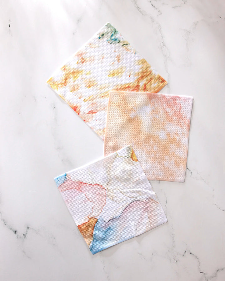 Three square cloths from GEOMETRY's Nice Water collection—featuring pastel marble patterns in yellow, orange, and multicolor—are displayed on white marble. Crafted from recycled materials, these super-absorbent dishtowels are both stylish and eco-friendly.