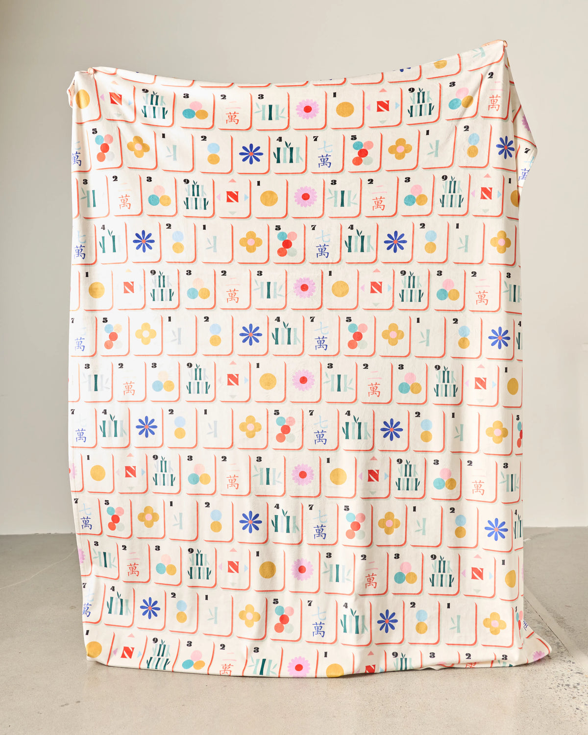 The Tile Toss Butter Blanket is made of light-colored fabric featuring a repeating grid of colorful mahjong tile illustrations.