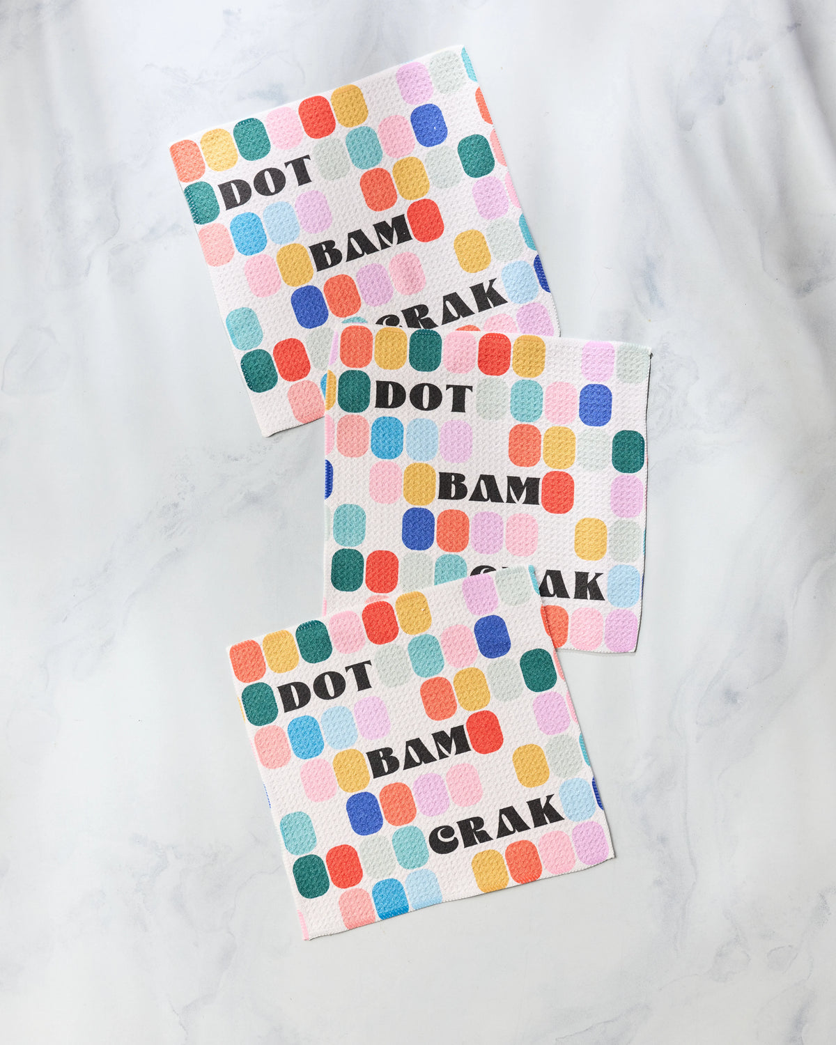 Three Geometry Tile Talk kitchen cleaning cloths stacked on a marble surface, featuring a colorful grid of rounded rectangles and the words DOT BAM CRAK in bold black letters.