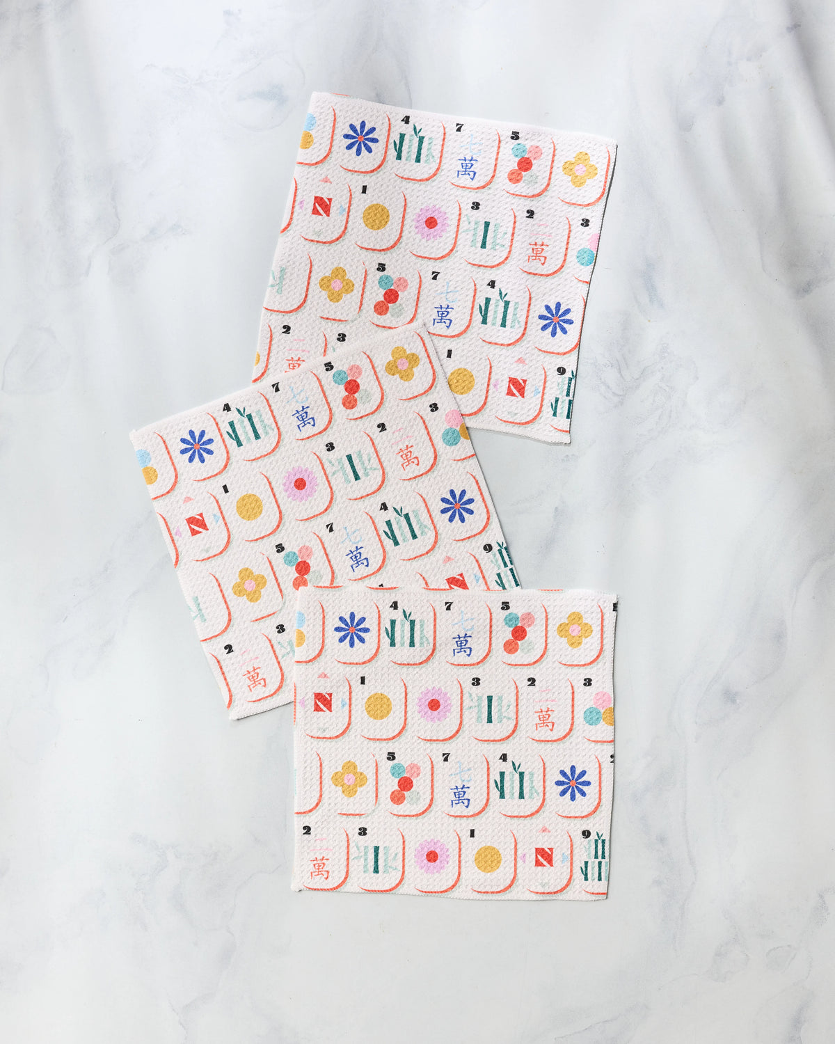 Three square, super-absorbent Geometry Tile Toss dishcloths featuring a colorful mahjong tile print laid out on a white marble surface.