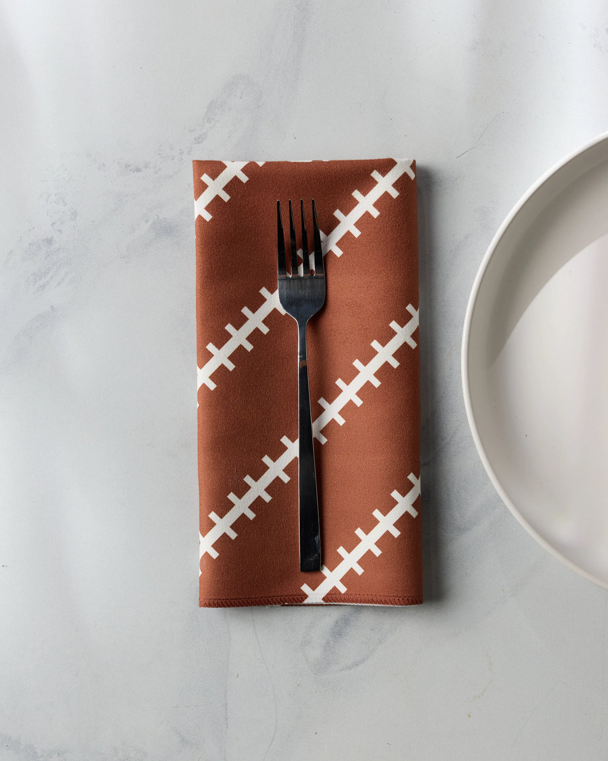 A brown Geometry Game Laces reusable table napkin with a white football lace pattern, topped with a silver fork, next to a white plate.