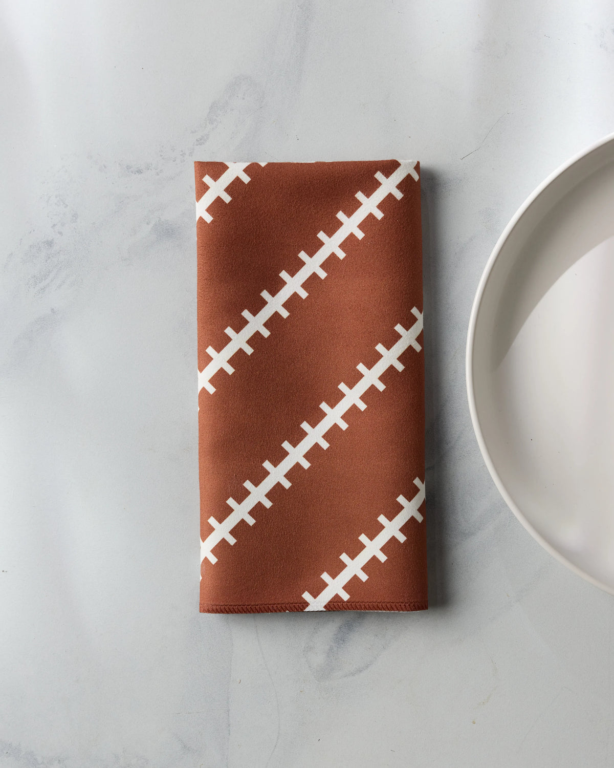 A brown Geometry Game Laces reusable table napkin featuring a white football lace pattern resting next to a white plate.