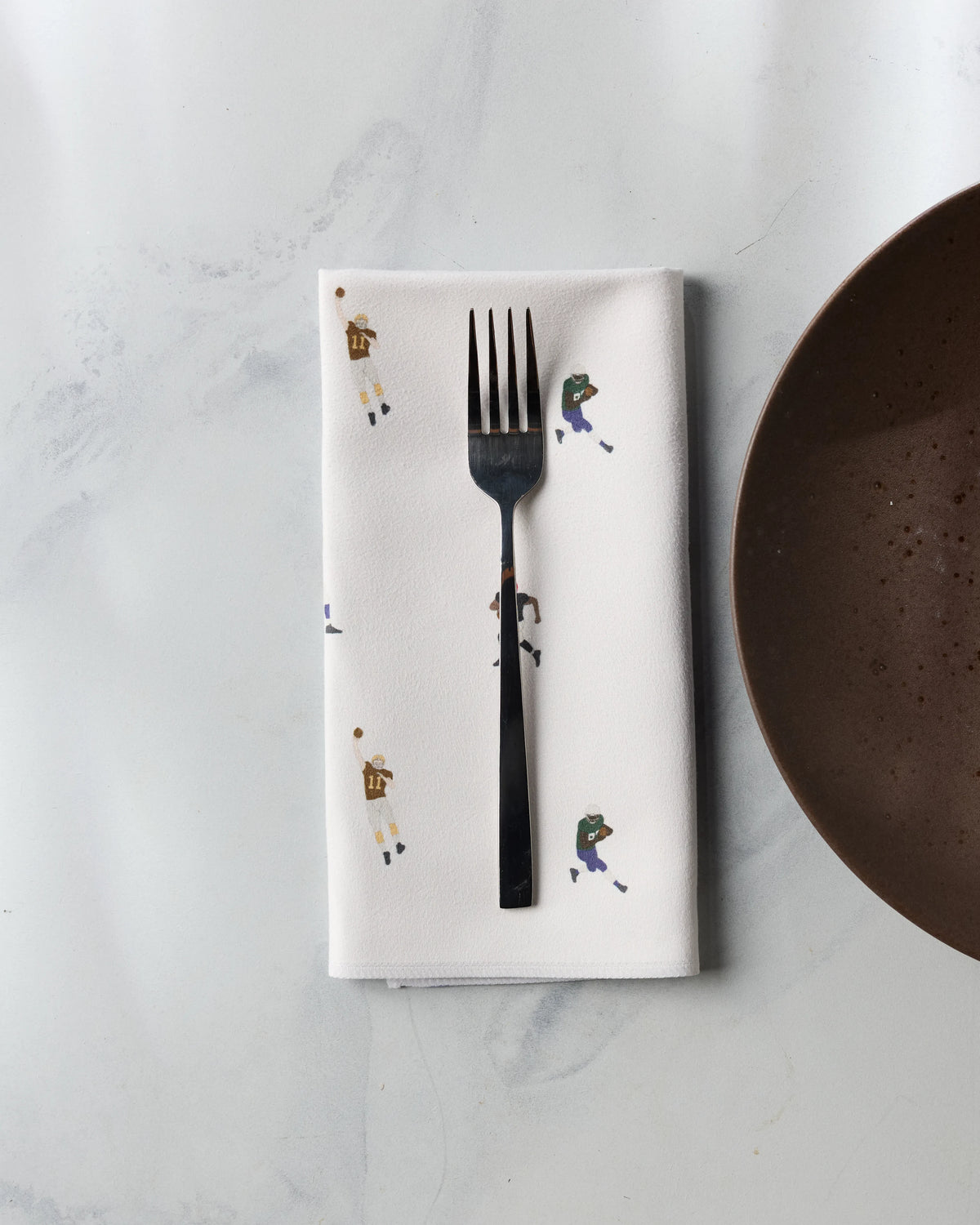 A folded white Geometry Roster dinner napkin patterned with small football players, topped with a stainless steel fork, beside a brown plate.