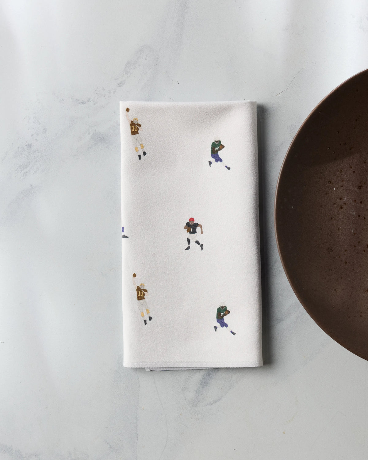 A folded white Geometry Roster dinner napkin with a pattern of small football players rests next to a dark brown plate.