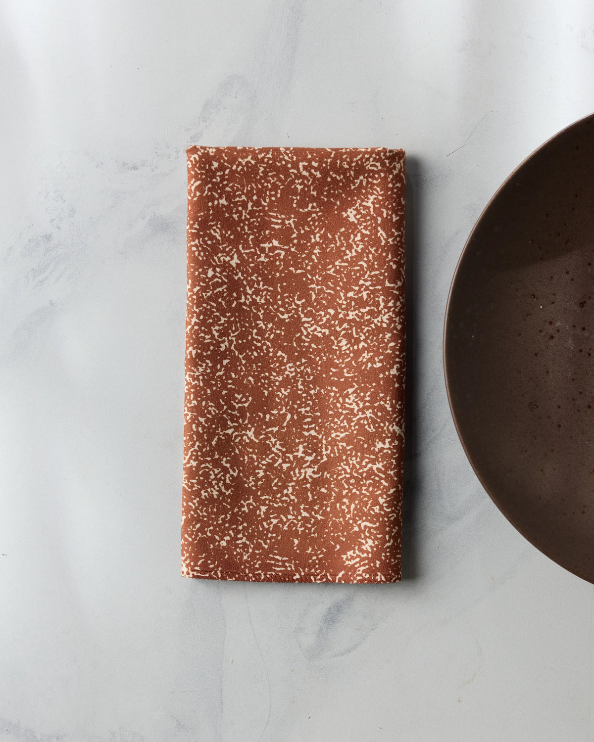 A folded rectangular Tonal Pebble dinner napkin with a rust-colored speckled pattern next to a brown plate.