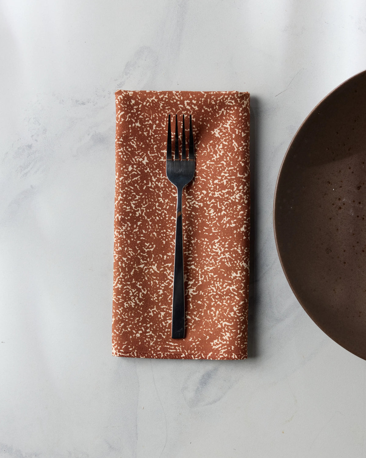 A metal fork resting on a rust-colored Geometry Tonal Pebble napkin with a white speckled pattern, placed next to a dark brown plate on a white marble surface.