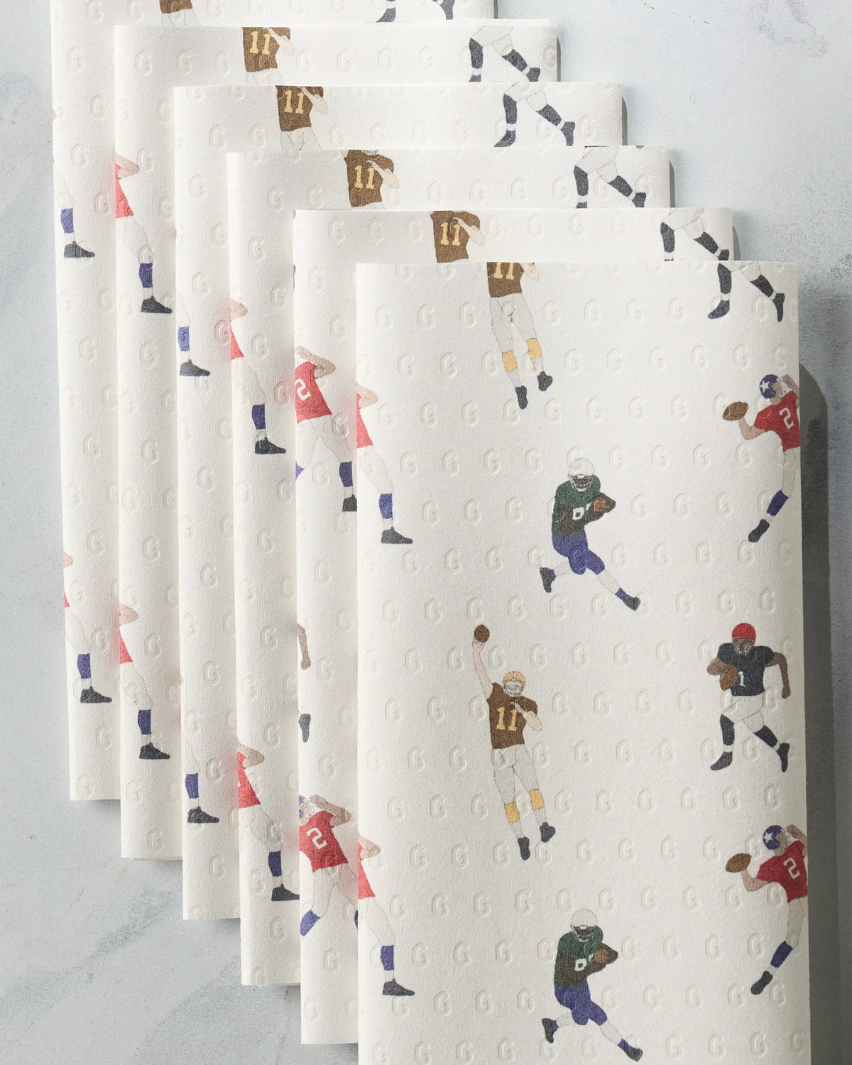 Five folded Geometry Roster reusable paper towels fanned out, each featuring a repeating pattern of football players in various uniforms against a light background embossed with a subtle G logo.