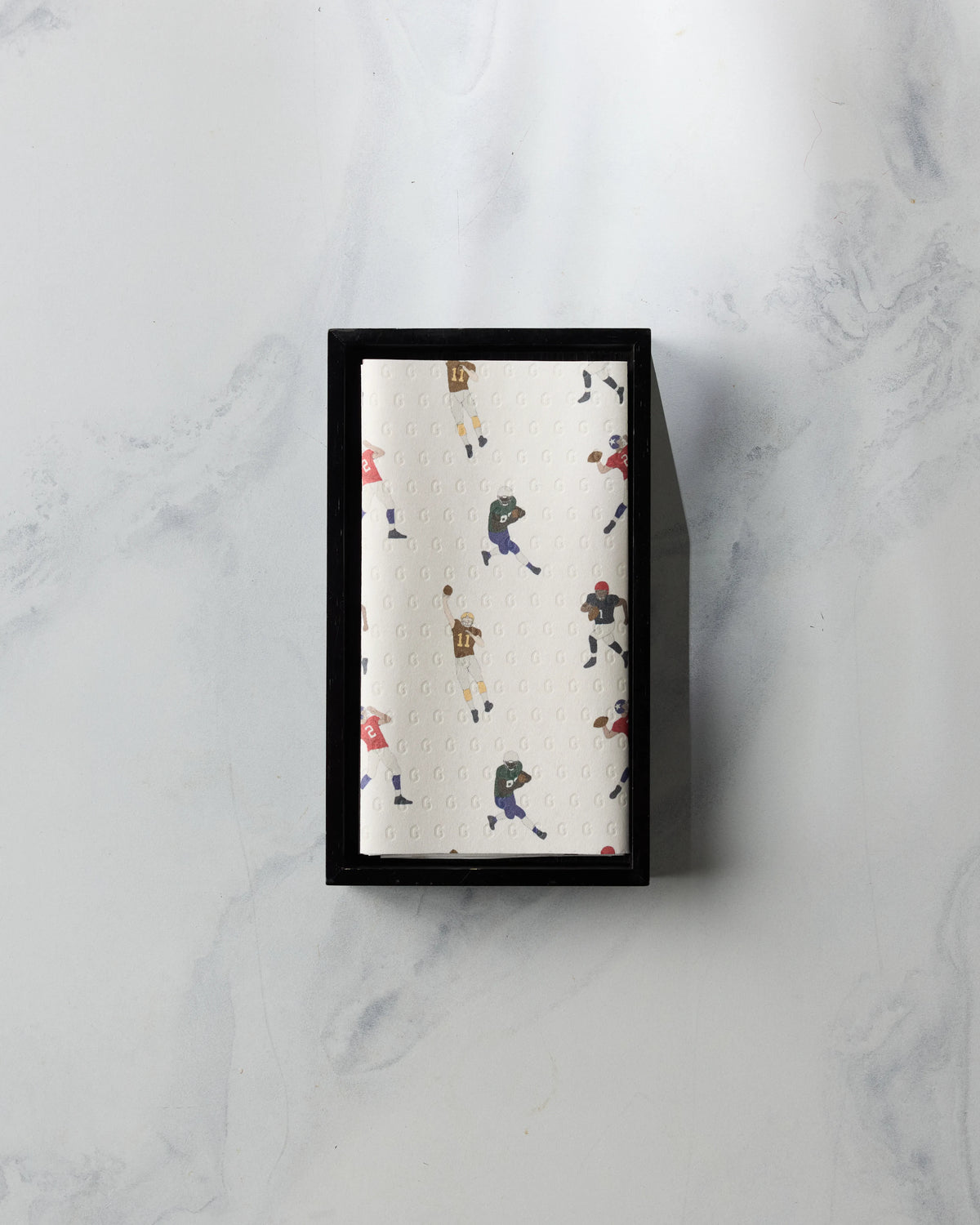 A folded white Geometry Roster towel printed with a pattern of football players rests in a black rectangular tray.
