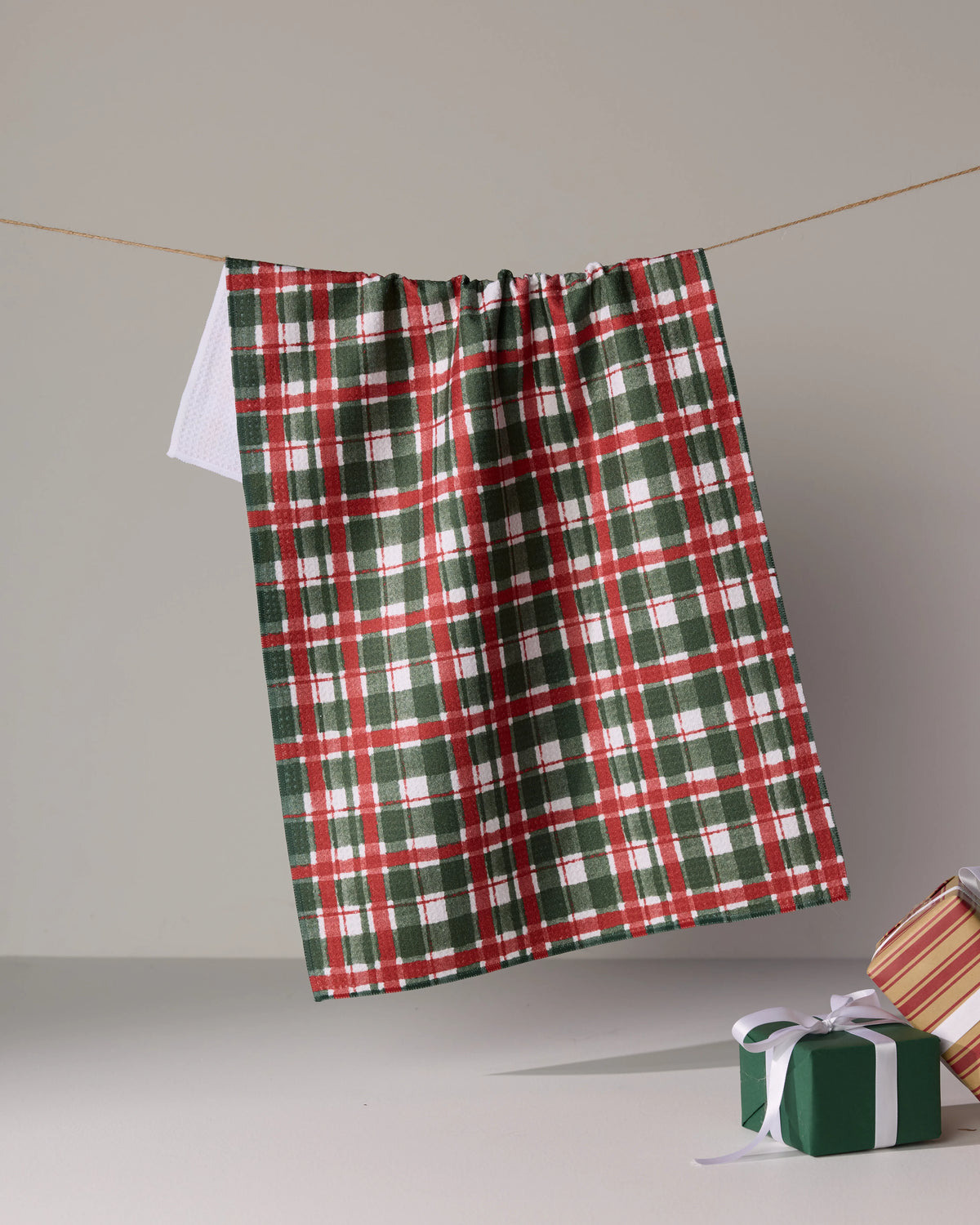A green, red, and white Alpine Plaid dish towel by Geometry hangs from a string. Two ribbon-wrapped gifts are partially visible in the lower right against a plain background.