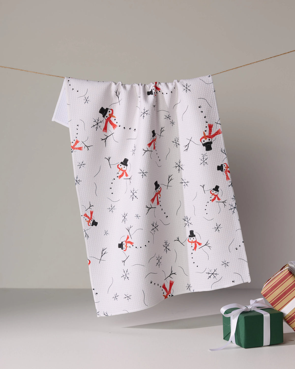 The Geometry Chill Chums white tea towel with red and black snowman print hangs on a line; next to it, two wrapped gifts—one green with a white ribbon and one striped—rest on a neutral background.