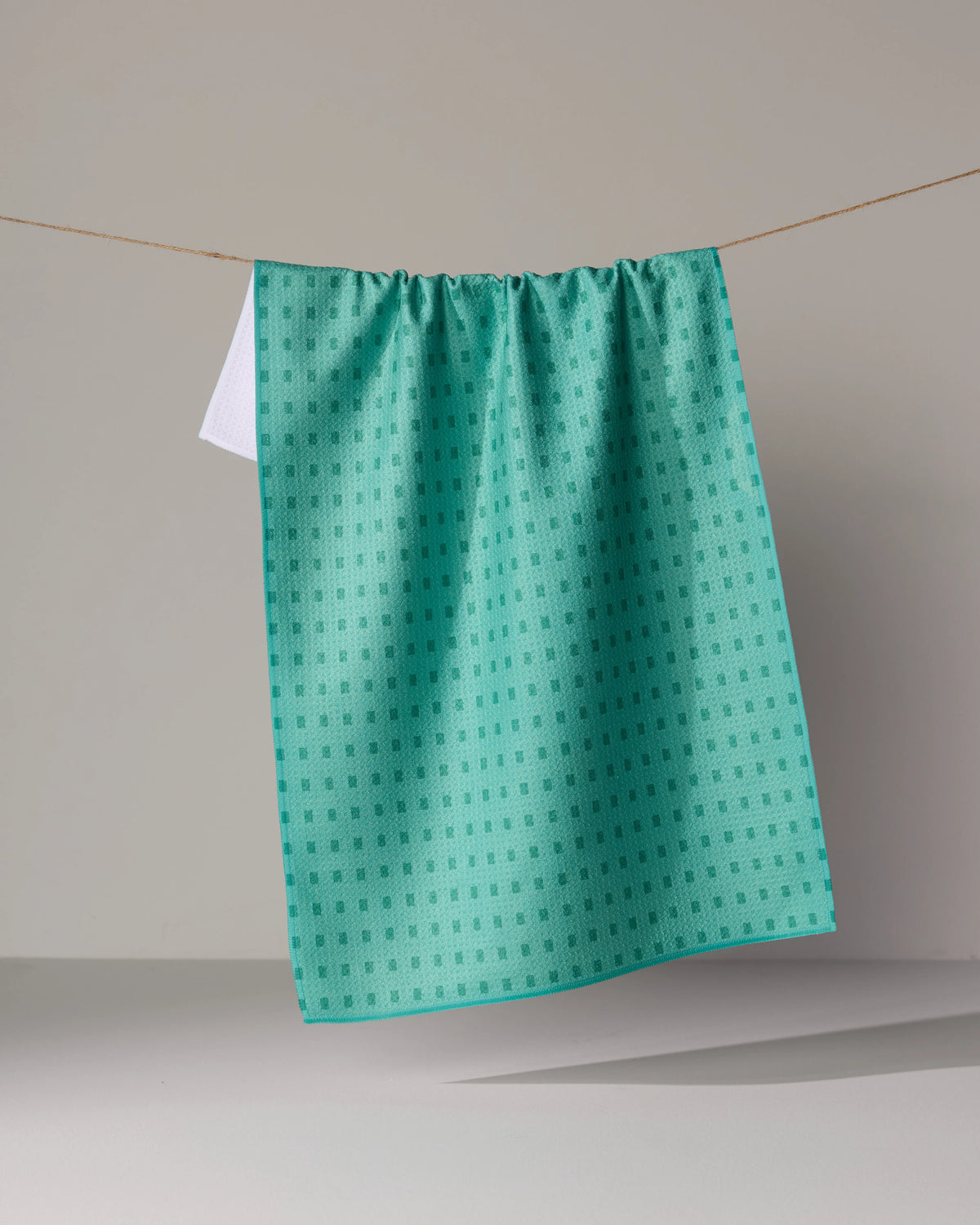 A teal Confetti Tiles tea towel with a repeating pattern of small squares hanging over a clothesline.