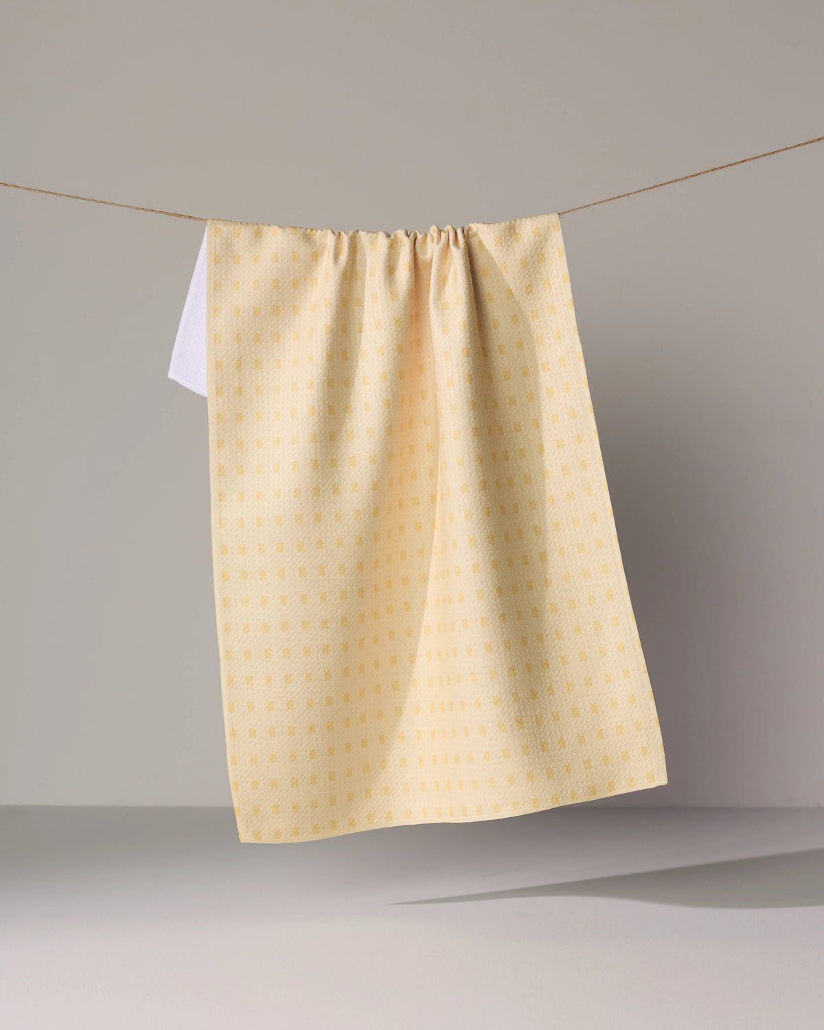 A yellow Geometry Confetti Tiles tea towel with a small repeating square pattern hanging from a string.