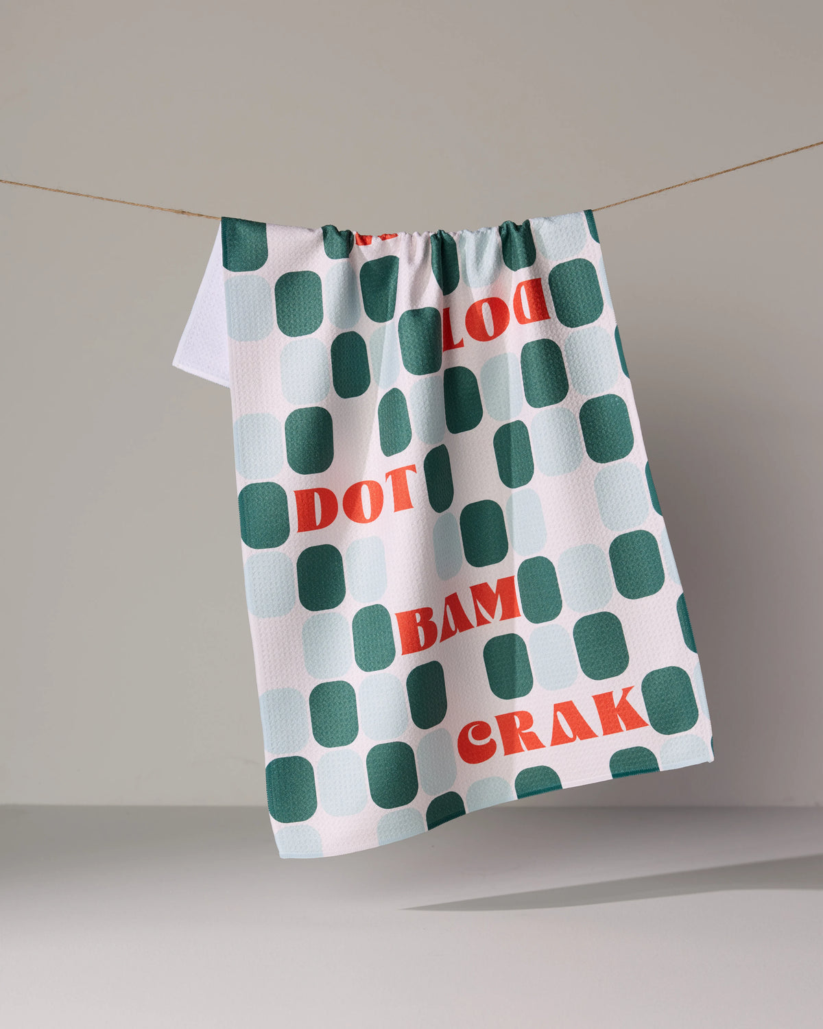 A Cypress Tile Talk recycled kitchen towel with a grid of green and light blue squares, featuring the red words DOT, BAM, and CRAK printed across it, hanging from a string.