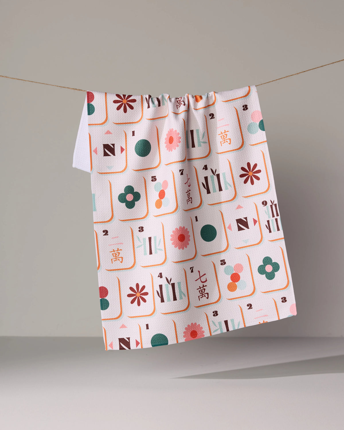 A Geometry Cypress Tile Toss quick-drying white tea towel featuring a repeating pattern of colorful mahjong tiles with floral and numerical designs hanging from a string.