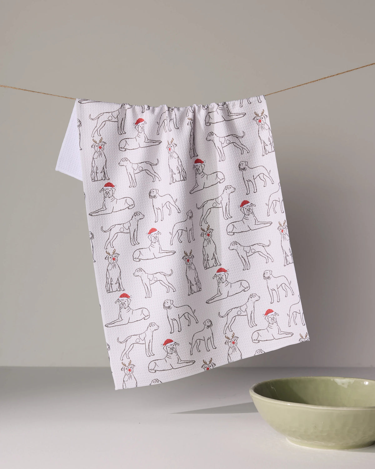 A white Geometry Festive Bulldogs tea towel with dog line drawings and red Santa hats hangs above a light green bowl on a plain surface.