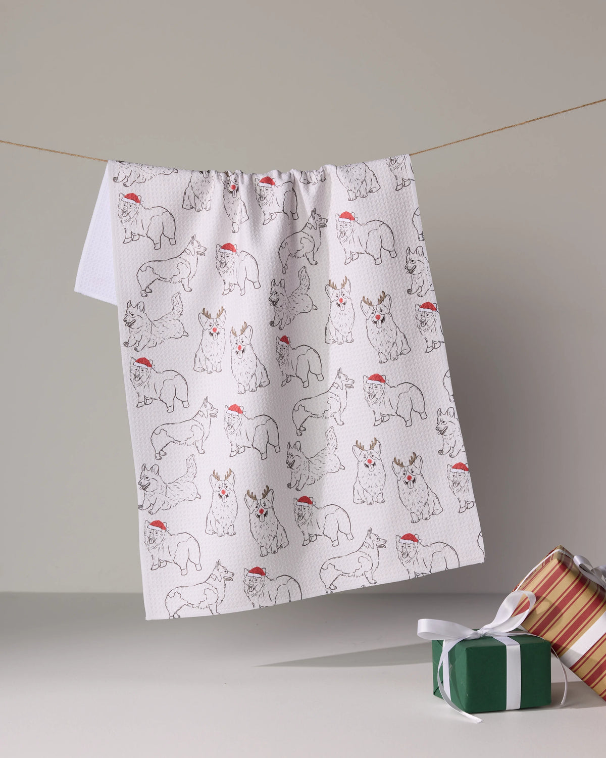The Geometry Festive Corgis tea towel, featuring white fabric with corgis in red Santa hats, hangs on a line beside two gift-wrapped presents set on a light surface.