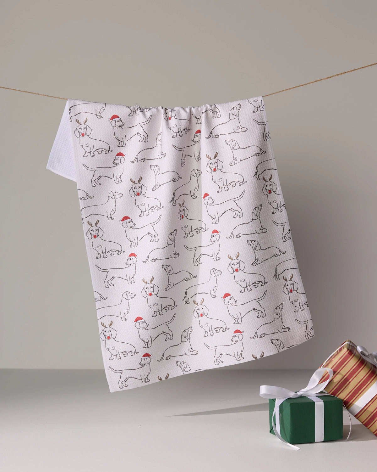 The Geometry Festive Doxies tea towel, made from quick-drying recycled materials, shows playful dogs in red Santa hats and hangs above two ribboned gifts.