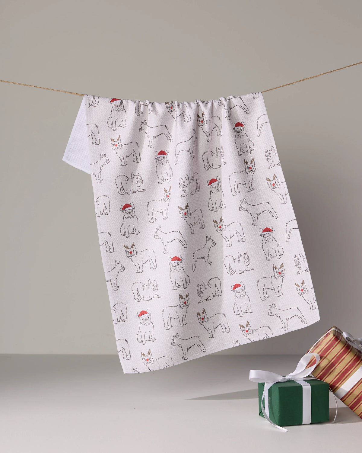 The Geometry Festive Frenchies quick-drying tea towel, made from recycled materials and featuring festive Frenchie illustrations, hangs above two holiday-wrapped presents.