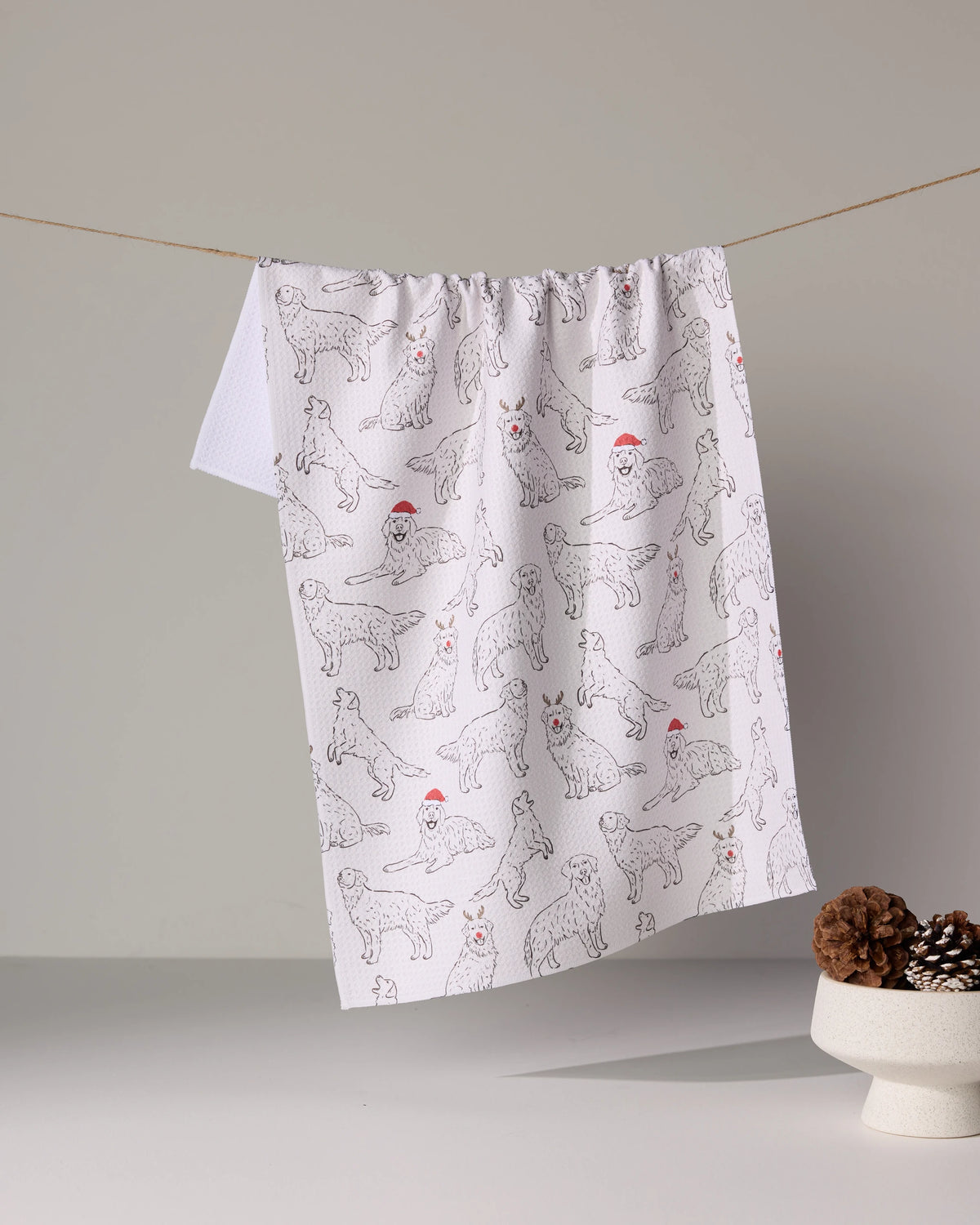 The Geometry Festive Goldens tea towel, featuring dog line drawings in red Santa hats, hangs on a string near pine cones. Made from quick-drying recycled materials, it’s set against a neutral background.