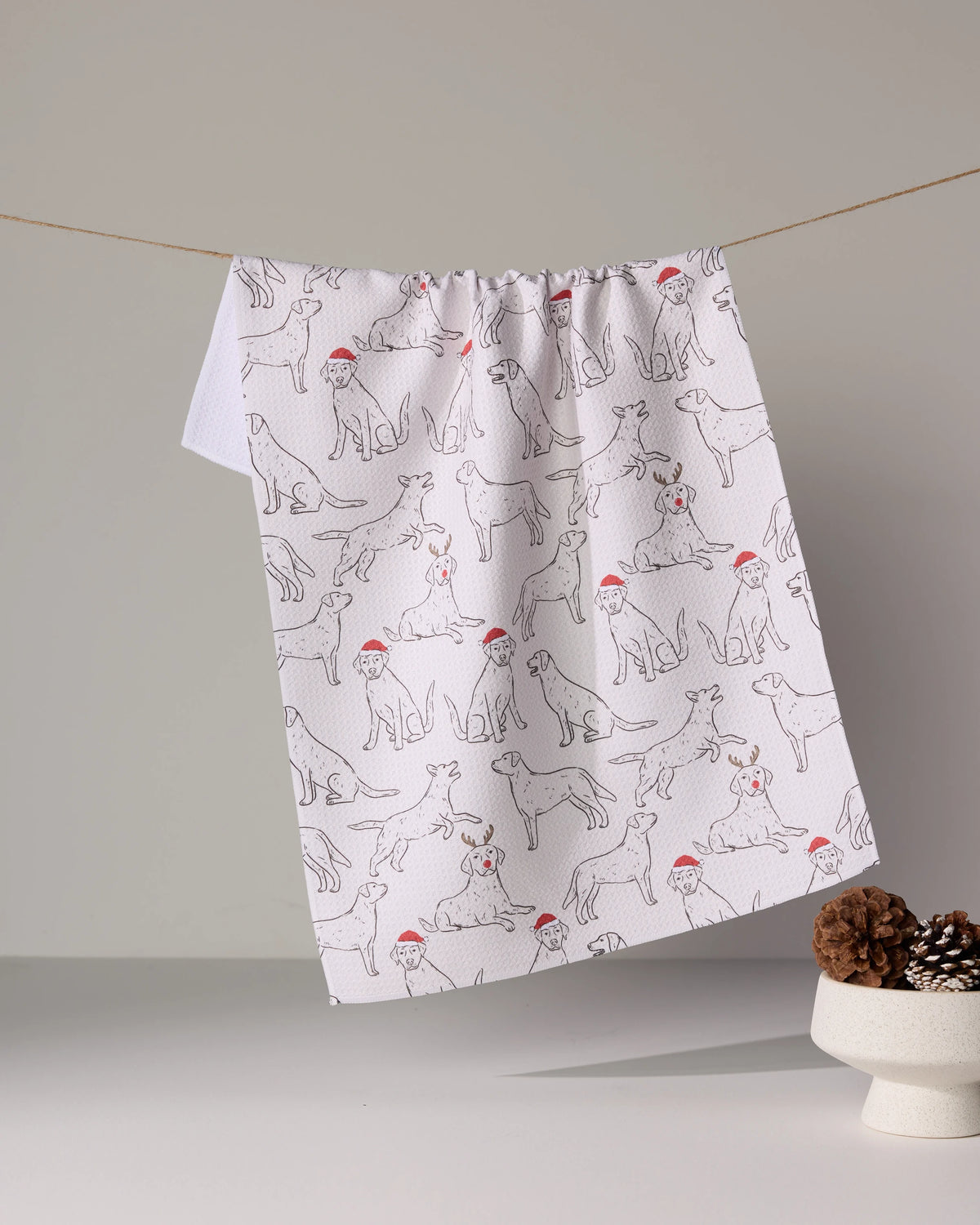 The Festive Labs tea towel by Geometry, featuring line-drawn dogs in red Santa hats, hangs on a string above a white surface with a pine cone-filled bowl in the foreground.