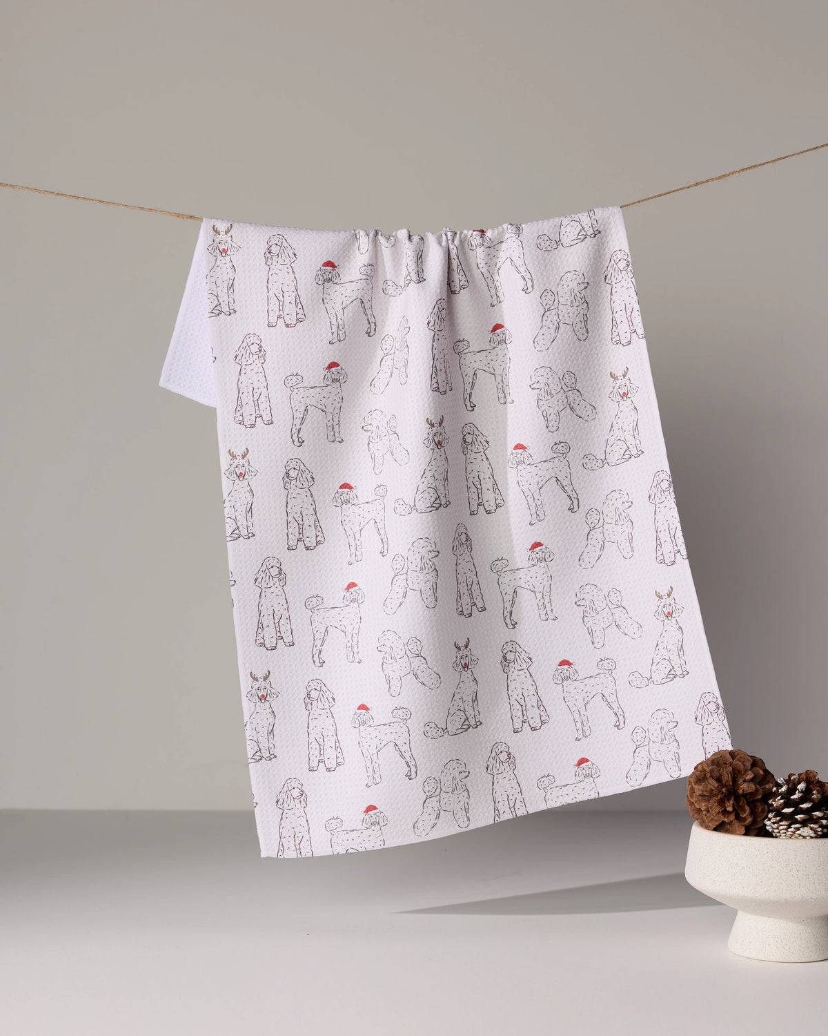 The Geometry Festive Poodles quick-dry tea towel, featuring poodles in red Santa hats, hangs on a string beside a white bowl of pinecones against a minimal backdrop.