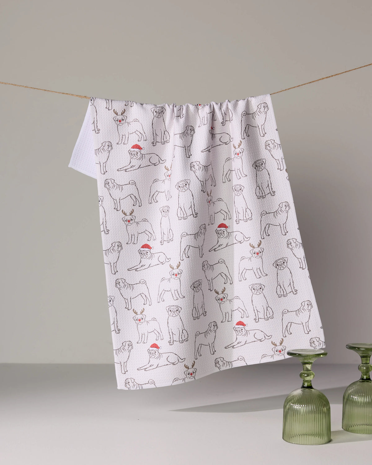 The Geometry Festive Pugs white kitchen towel with outlined dogs in Santa hats and scarves, made from recycled materials, hangs on a clothesline near two green glass cups on a gray surface.
