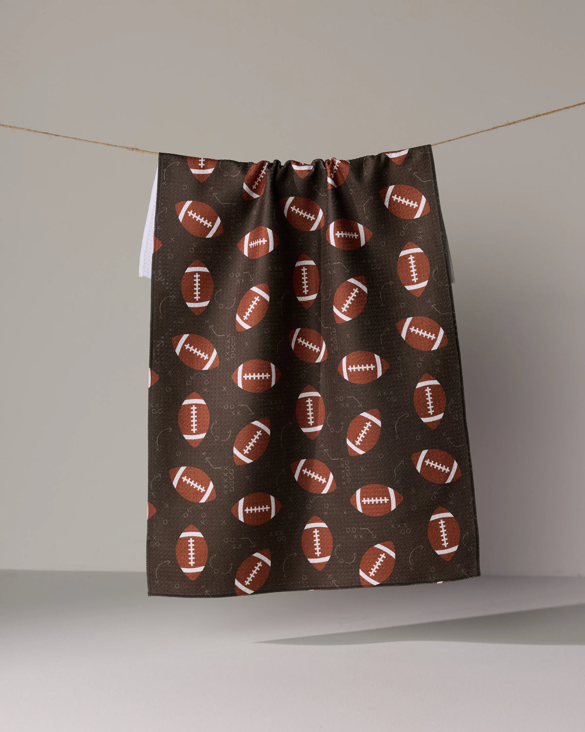 Geometry First Down dark brown quick-drying tea towel with a repeating pattern of footballs and white line play diagrams hanging on a string.