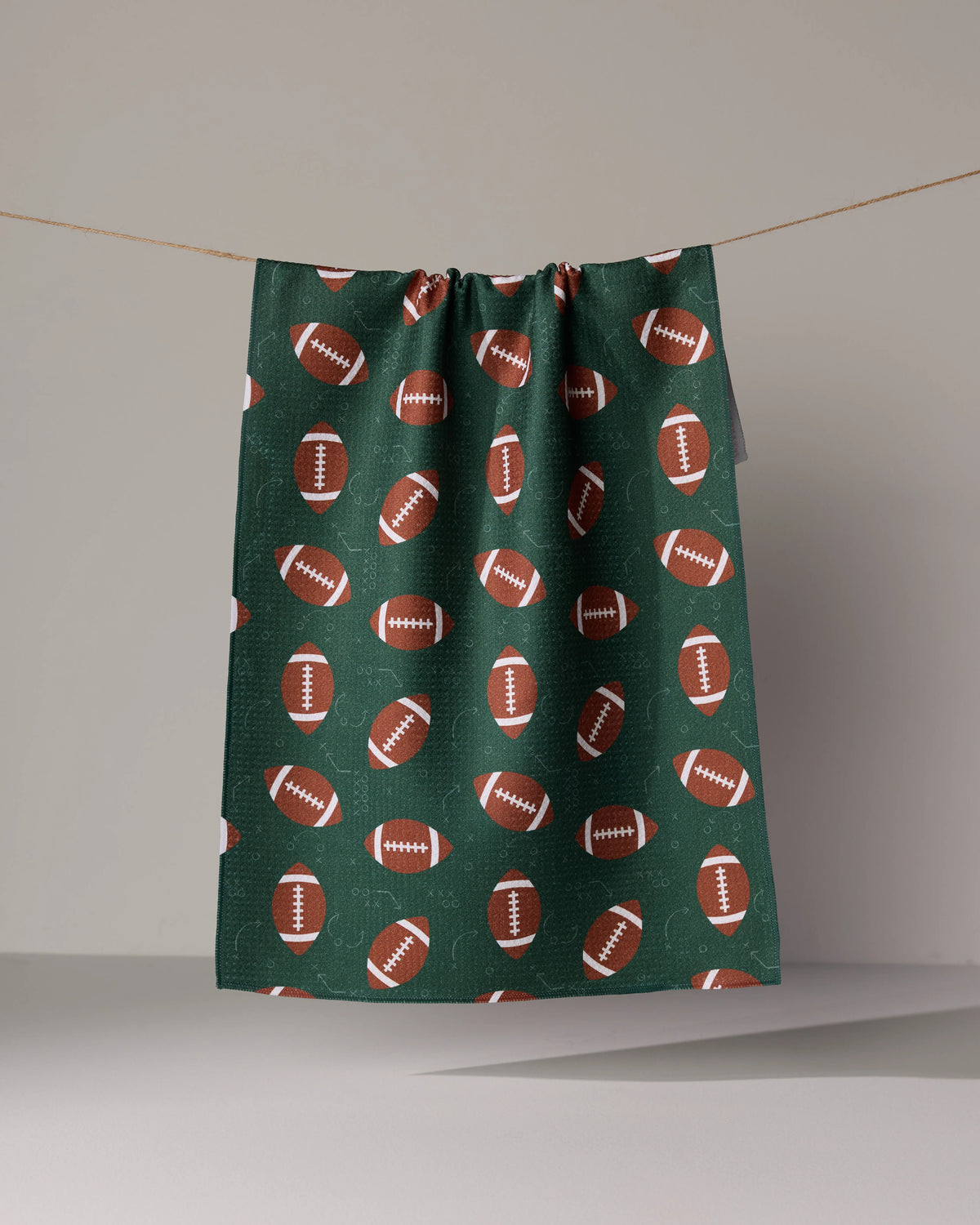 A Geometry First Down tea towel in dark green with brown footballs and faint white play diagrams hanging on a clothesline.