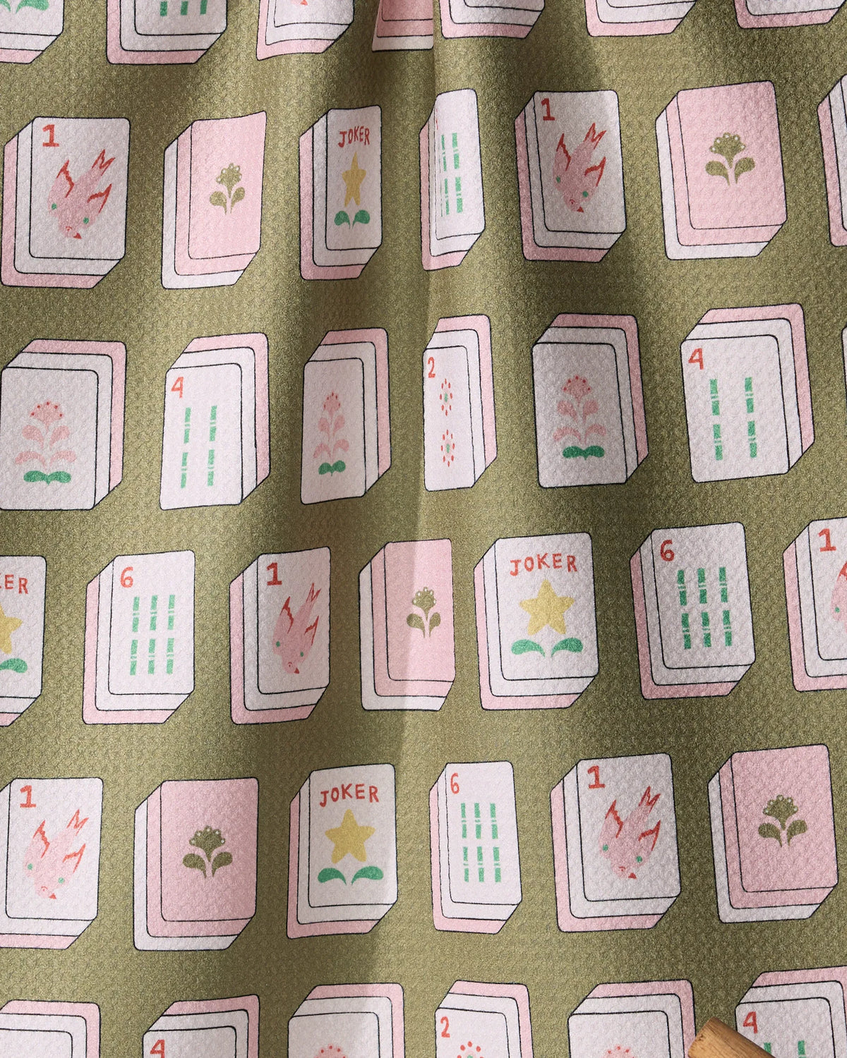 A Geometry Flower Tiles tea towel featuring pale pink and white mahjong tiles decorated with flowers, numbers, and star symbols on an olive green background.