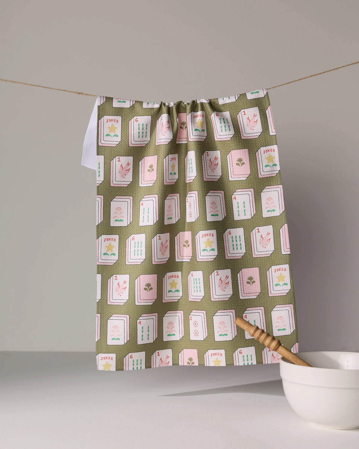 A Flower Tiles kitchen towel made from recycled materials, featuring a repeating pattern of mahjong tiles on a green background, hanging from a clothesline.