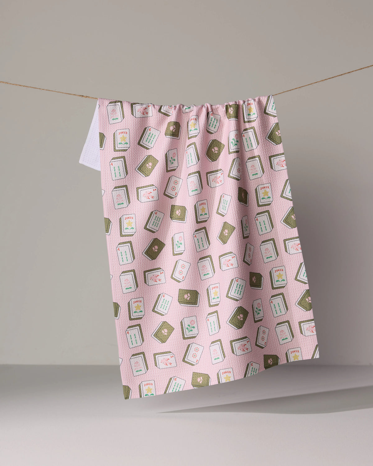 A pink waffle-weave Flower Tiles tea towel patterned with illustrations of mahjong tiles hanging on a clothesline.
