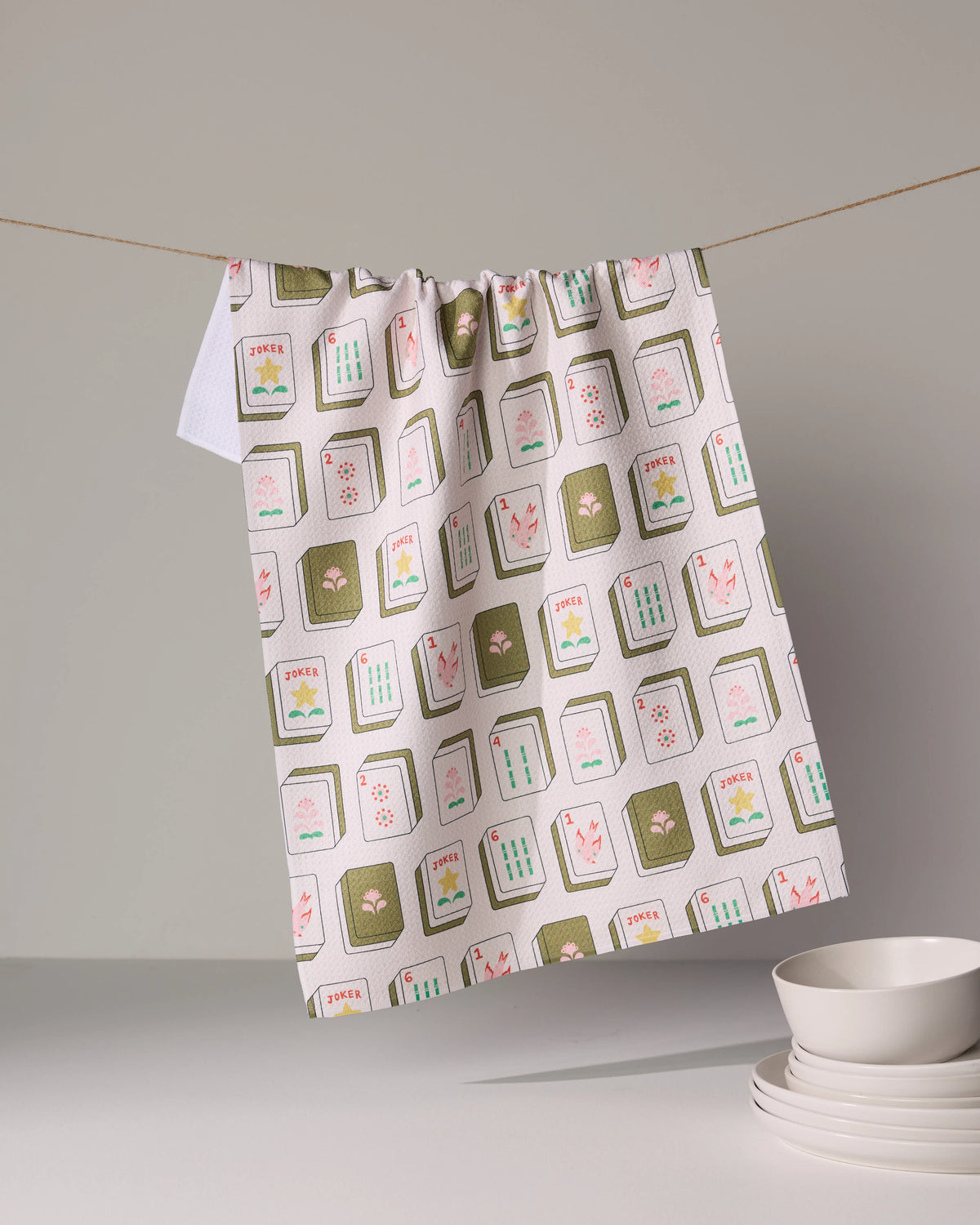 A Flower Tiles patterned tea towel by Geometry hangs on a string above a stack of white bowls.