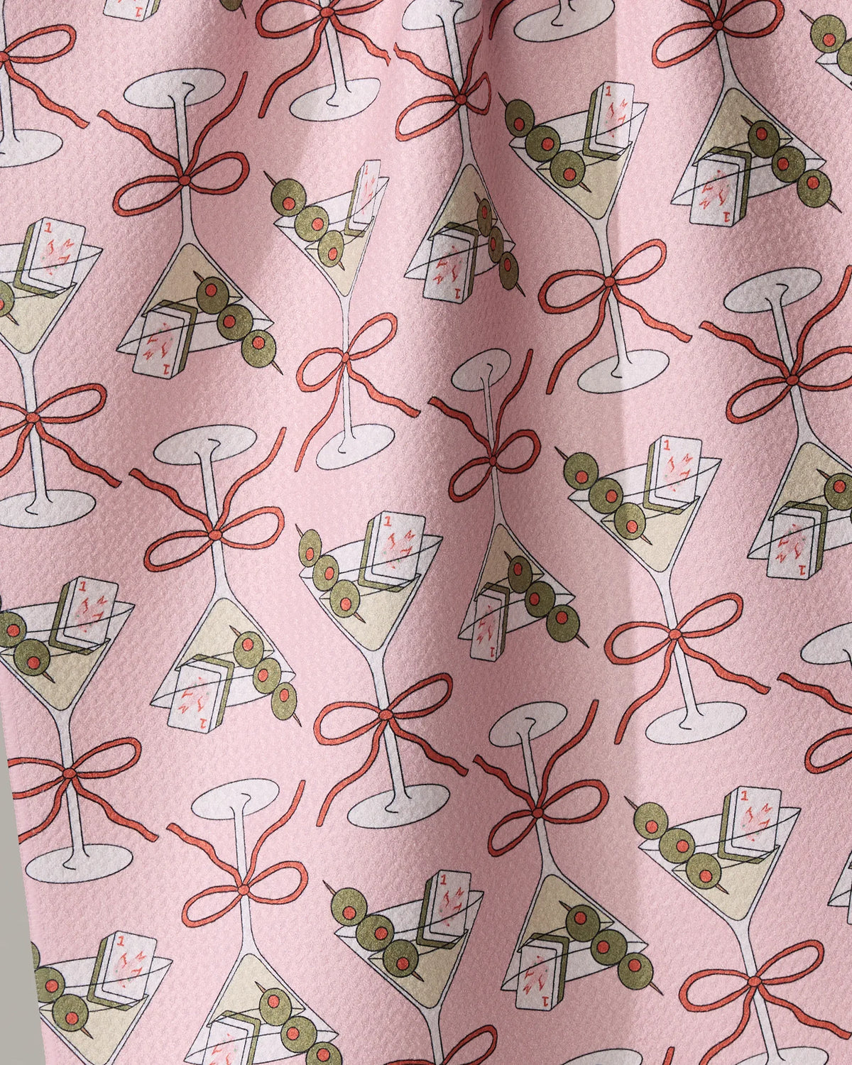 Geometry Flower-tinis tea towel featuring martini glasses, each garnished with three olives and accompanied by a mahjong tile, tied with a red ribbon on a pink background.