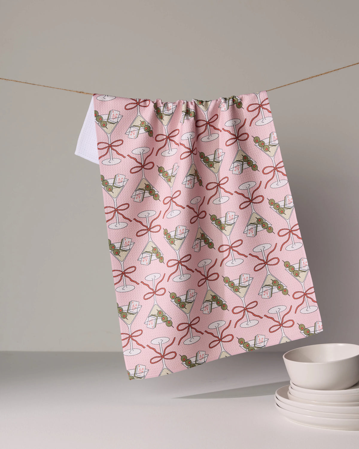 A pink Geometry Flower-tinis recycled kitchen towel with a repeating pattern of martini glasses, red ribbons, and olives, hanging on a string above a stack of bowls.