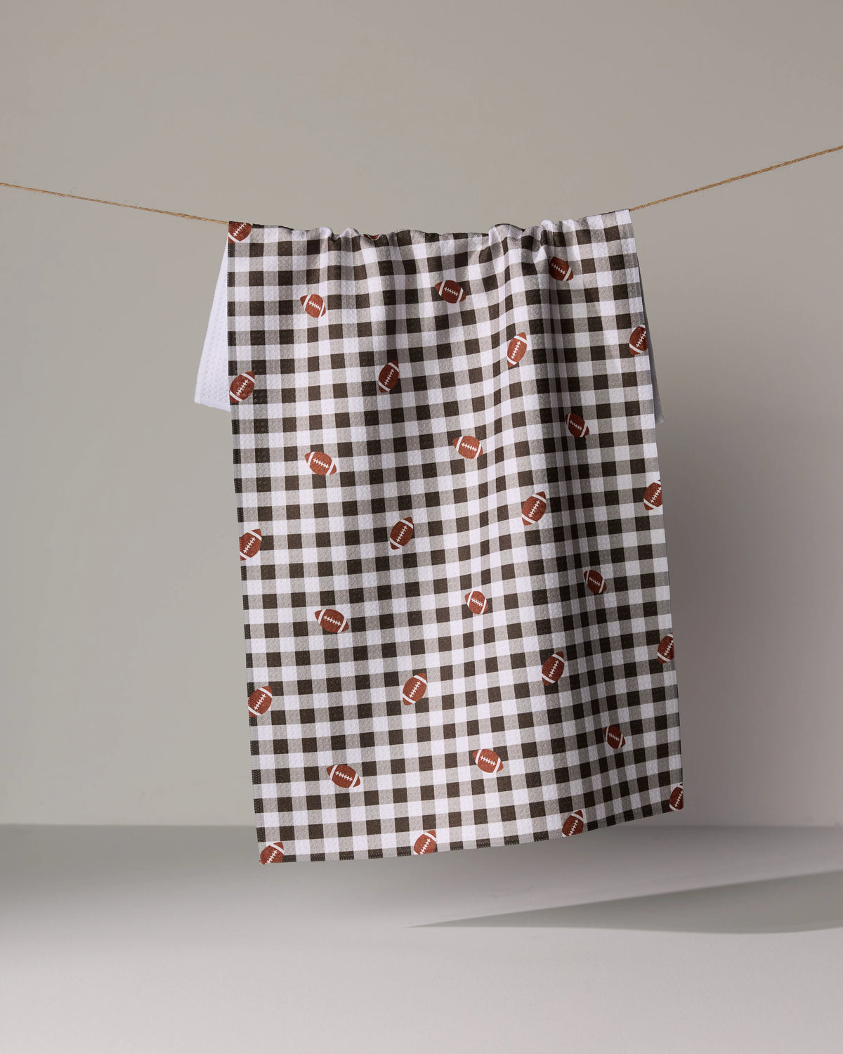 A brown and white Football Check kitchen towel decorated with small football icons hanging on a string.