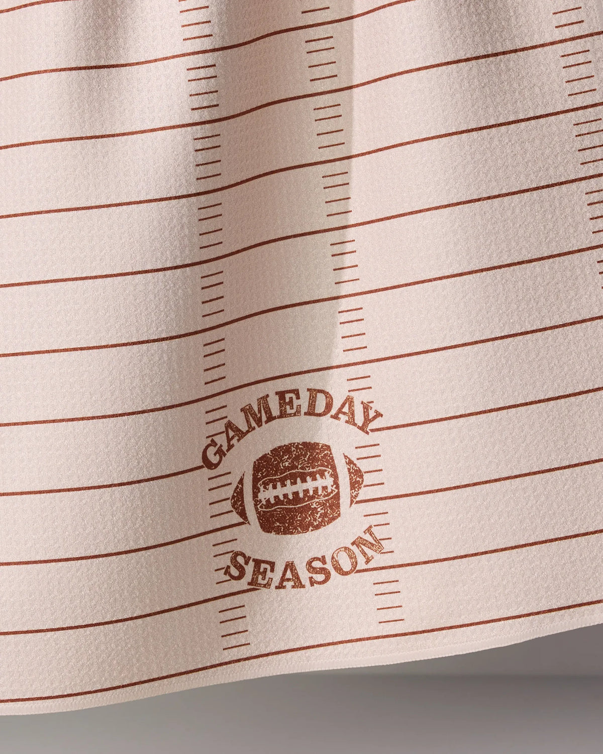 Off-white Geometry Gameday Season tea towel with horizontal brown lines resembling a football field, featuring a football graphic and the text GAMEDAY SEASON in the center.