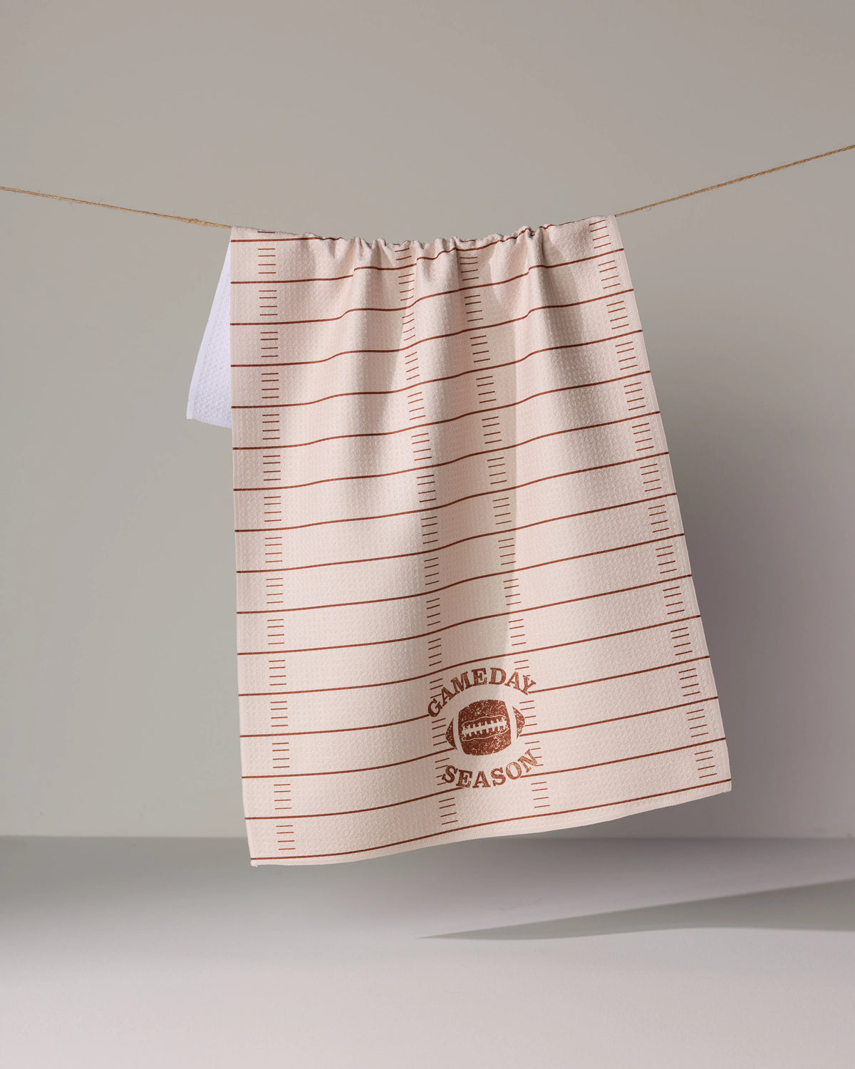 A Gameday Season Geoweave tea towel with a football field pattern and a football graphic, hanging on a clothesline.