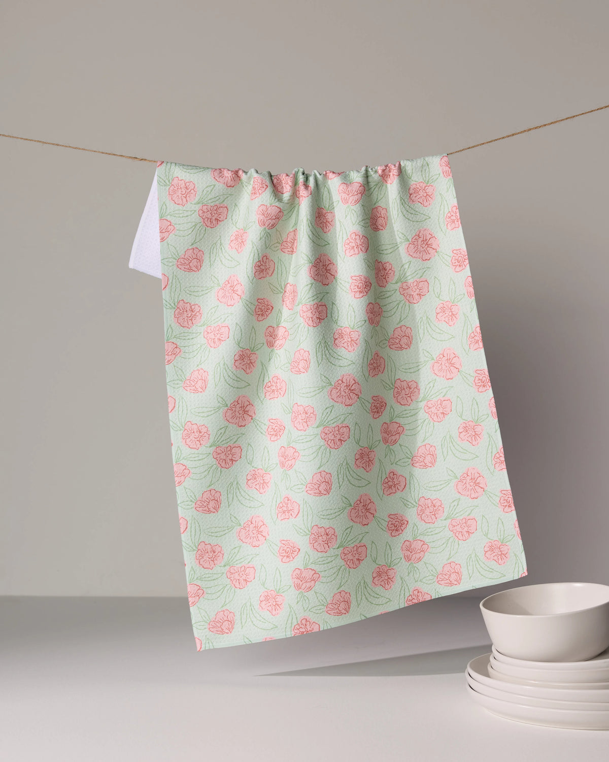 A Geometry Lucky Bloom floral tea towel patterned with pink flowers and green leaves hangs on a line above a stack of bowls.