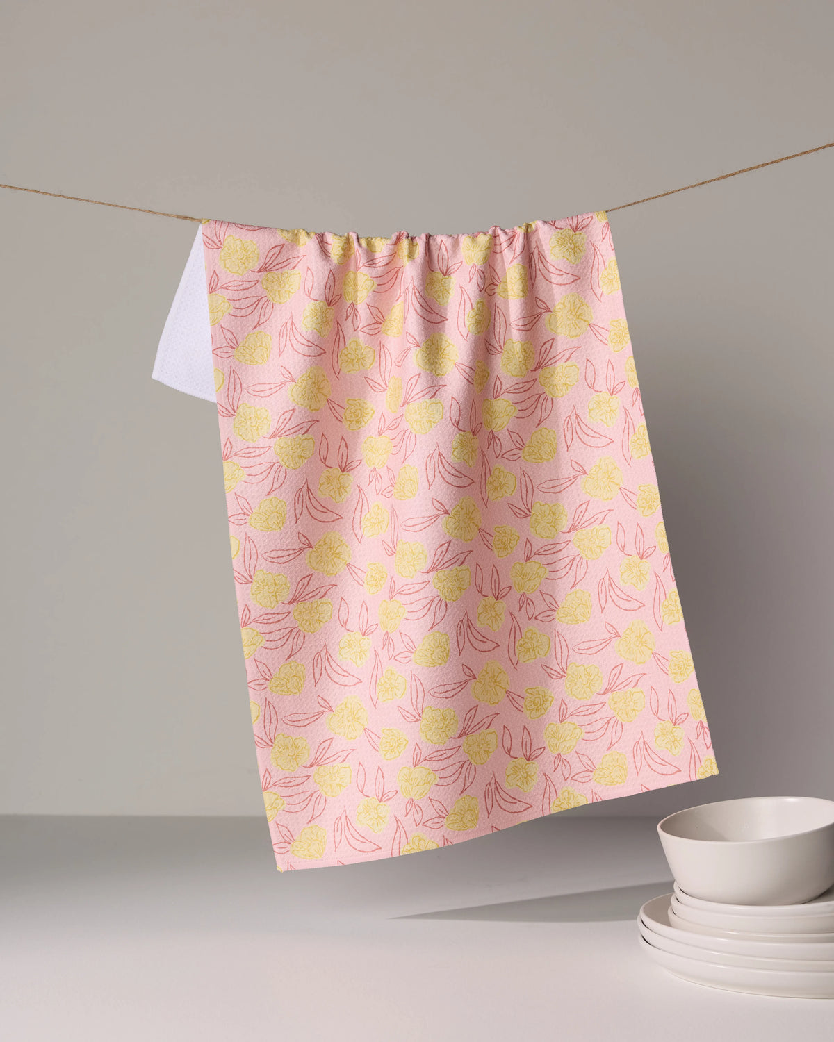 A pink floral Geometry Lucky Bloom kitchen towel hangs on a string above a stack of white bowls.