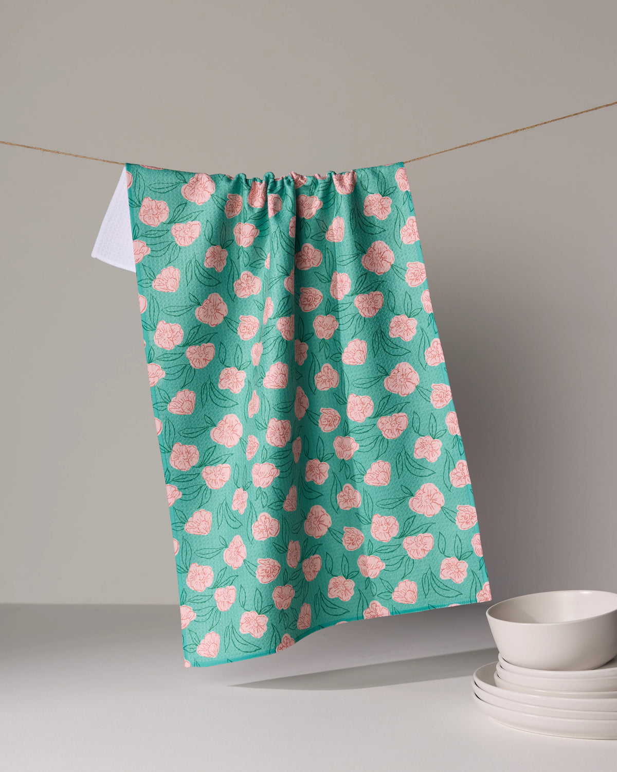Lucky Bloom teal kitchen towel with a pink floral pattern hanging from a clothesline above a stack of white bowls.