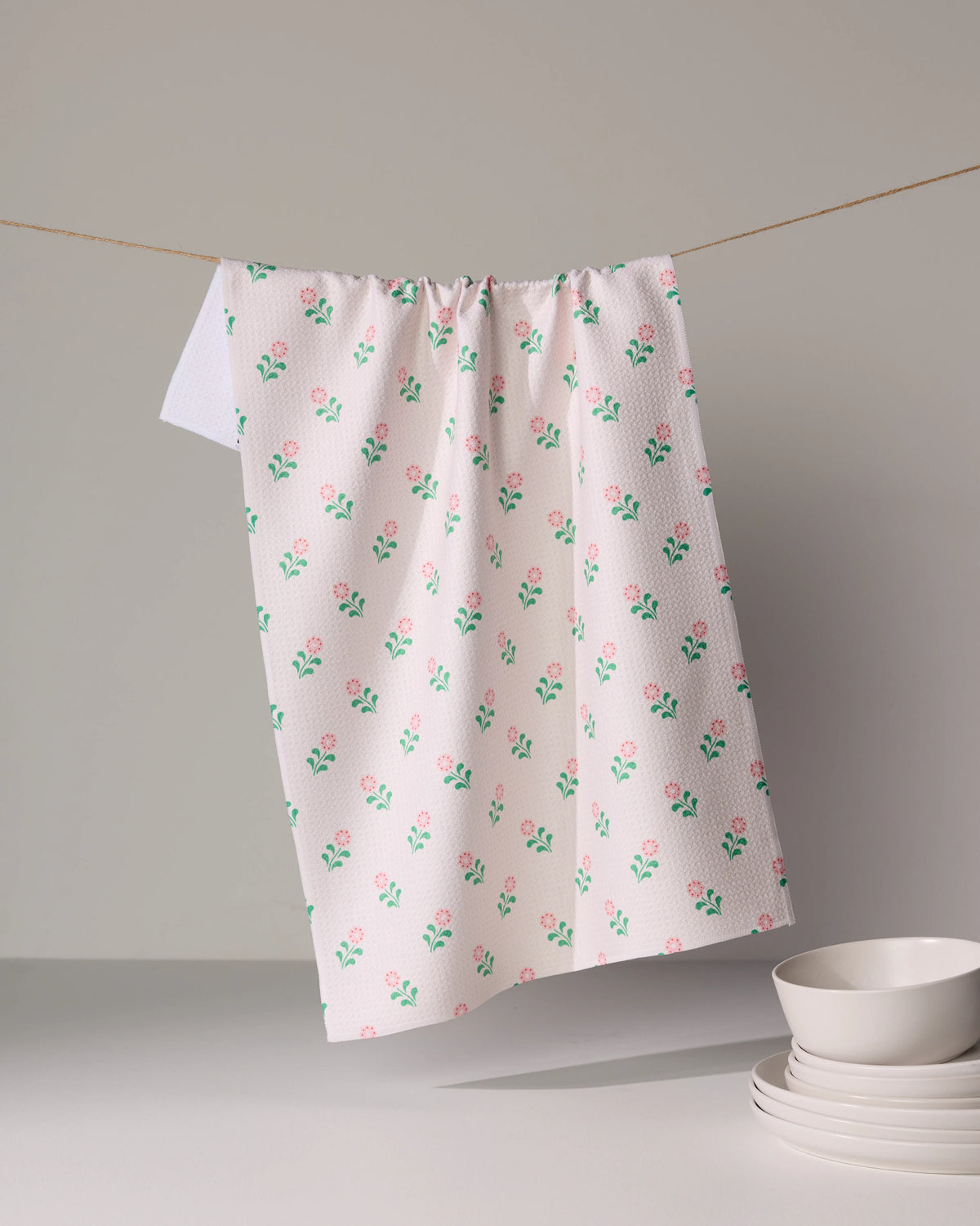A Geometry Lucky Blossom ultra-absorbent kitchen tea towel featuring small pink flowers and green stems draped over a string, with a stack of white bowls nearby.