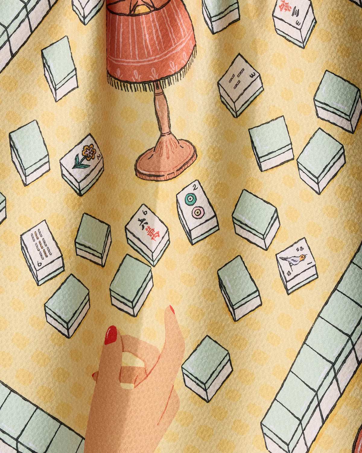 A hand reaches toward scattered mahjong tiles on a Lucky Hand tea towel by Geometry, which features a yellow pattern and is set beside a floor lamp.