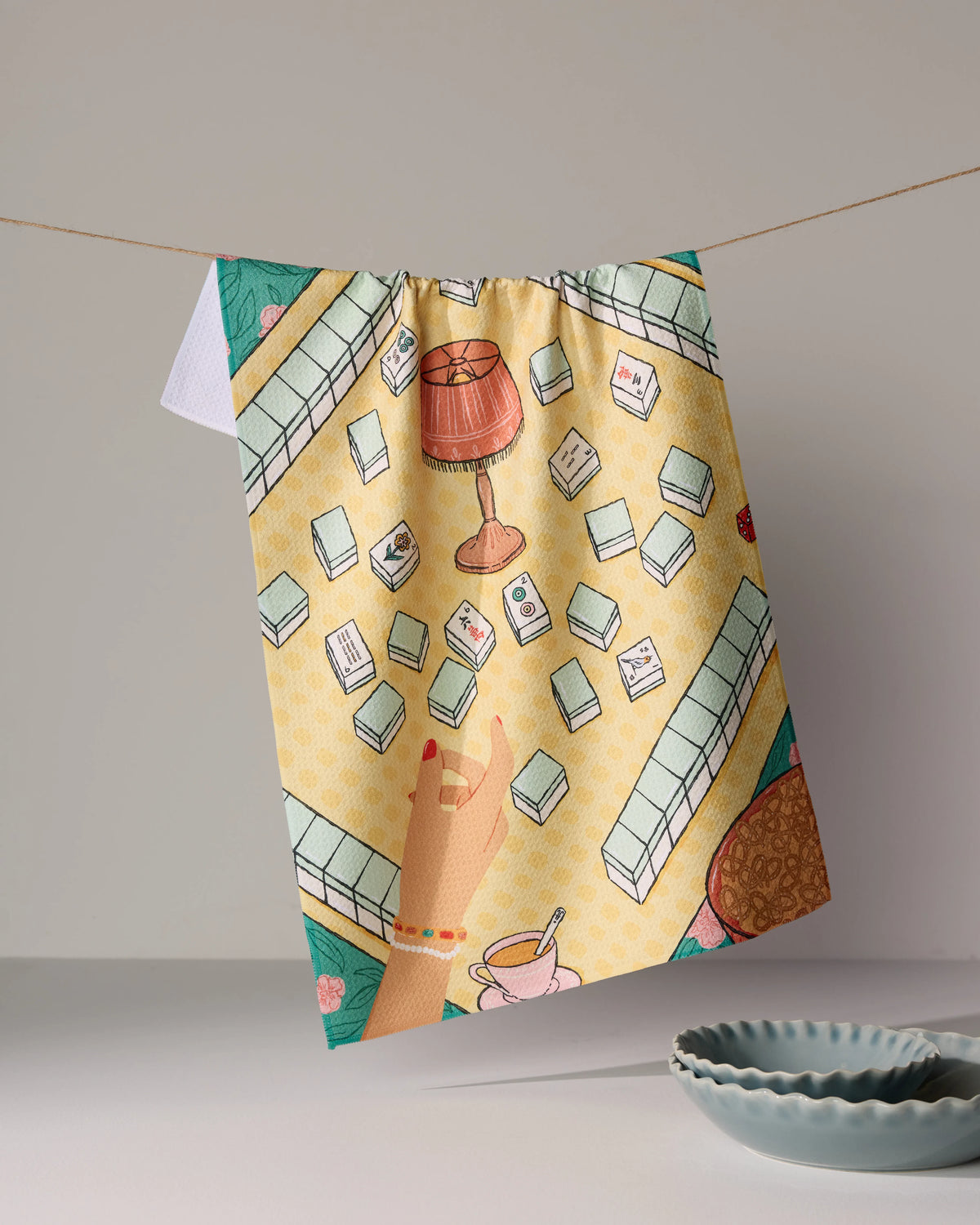 A Geometry Lucky Hand tea towel hangs on a string, featuring a patterned illustration of a Mahjong game in progress, including a hand reaching for a tile, a table lamp, and a teacup.